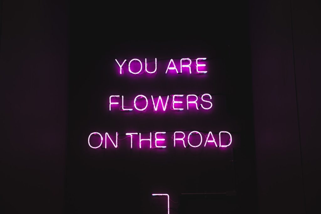 Pink color neon luminous text with inspiring phrase You are flowers on the road on black signage at night that says: "YOU ARE FLOWERS ON THE ROAD".