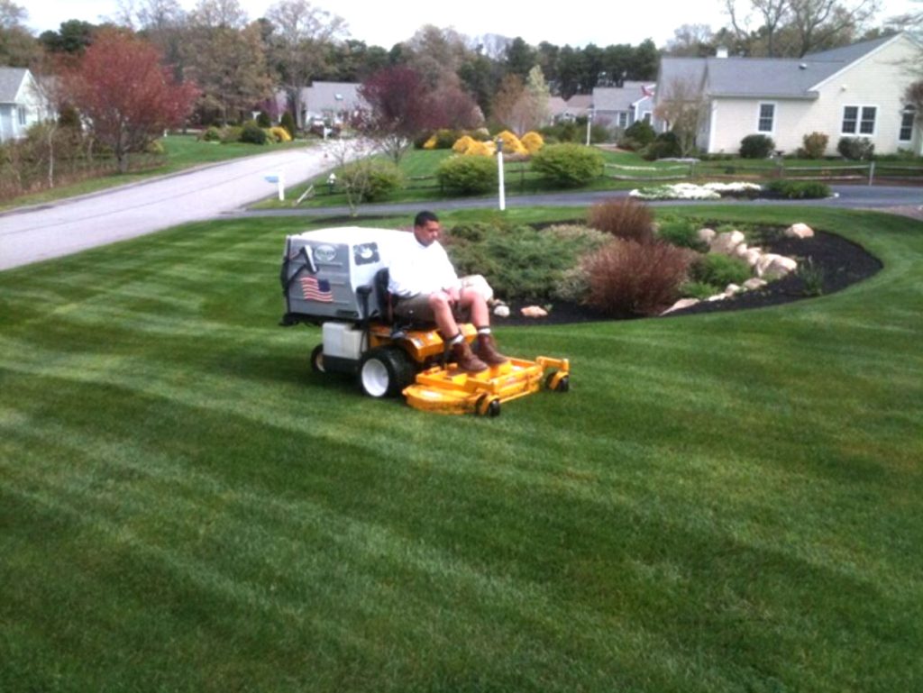 Man on yellow riding lawn mower with beautiful lawn and large house.