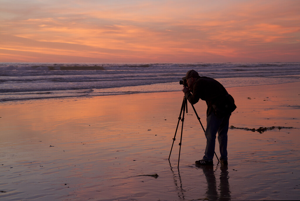 Photographer on a beach with tripod and a pink sky.